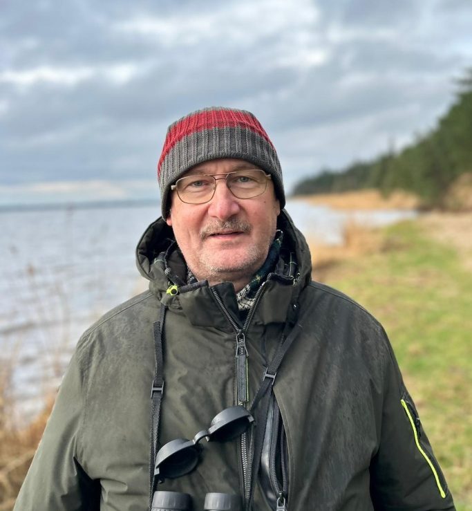 Mann mit Brille und Hut steht am Strand, im Hintergrund Wasser und Bäume.