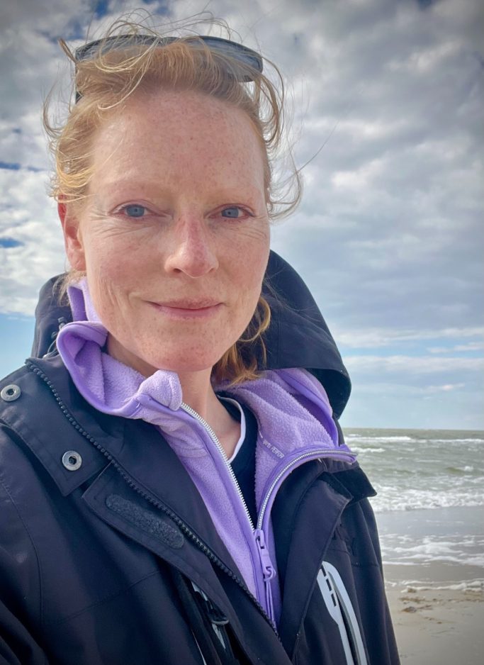 Frau mit roten Haaren, Steht am Strand, trägt einen schwarzen Anorak und schaut in die Kamera.