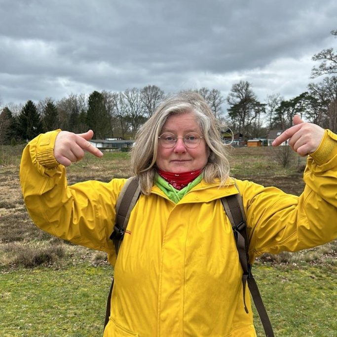 Frau mit Brille in gelbem Mantel zeigt mit beiden Händen auf sich in einem Feld.