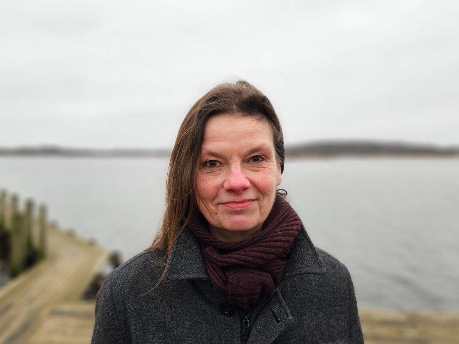 Frau mit braunen Haaren und Schal, vor einem Gewässer an einem bewölkten Tag.