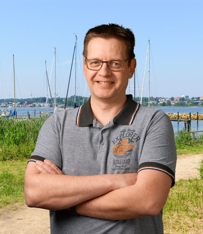 Mann mit Brille, lächelnd, steht am Ufer mit Segelbooten im Hintergrund.