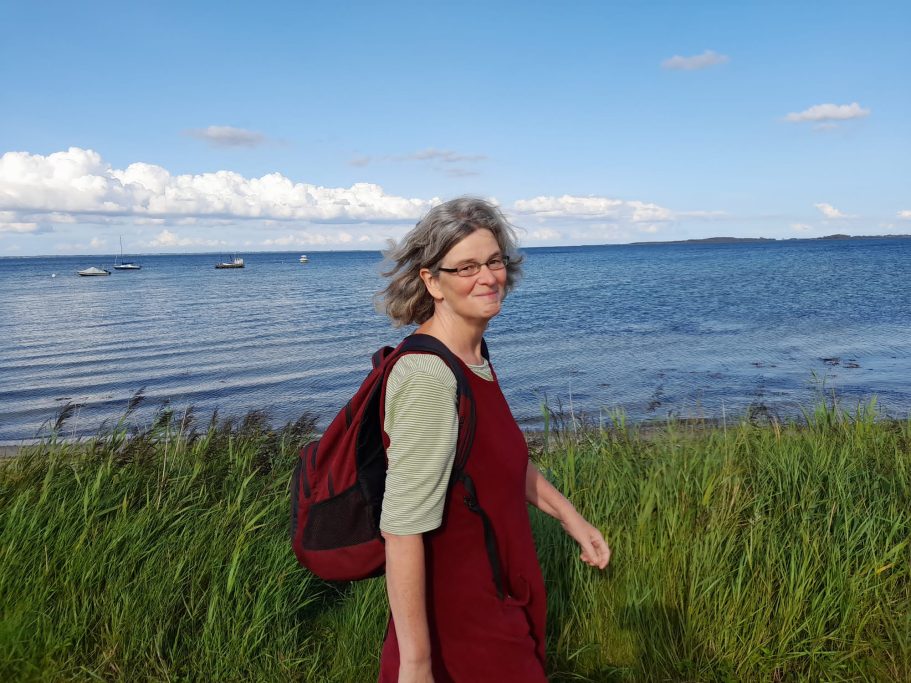 Frau mit grauen Haaren und Rucksack steht am Ufer eines Gewässers, umgeben von Gras.