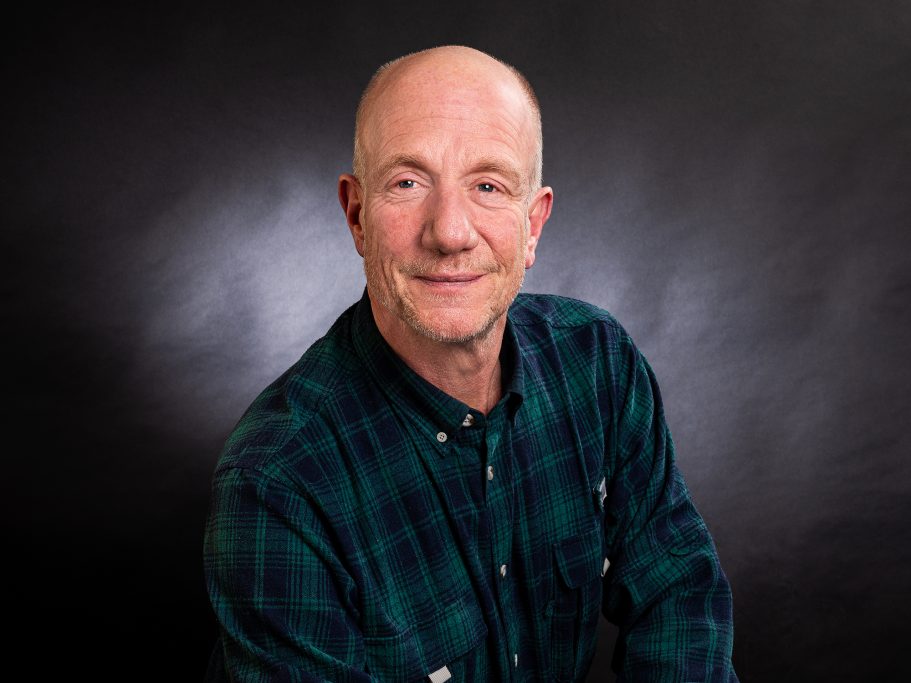 Mann mit Glatze und freundlichem Lächeln, trägt ein kariertes Hemd.