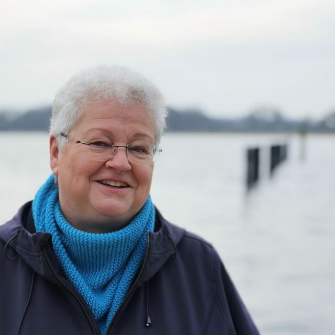 Lächelnde ältere Frau mit Kurzhaarschnitt steht am Wasser, im Hintergrund Pfähle.