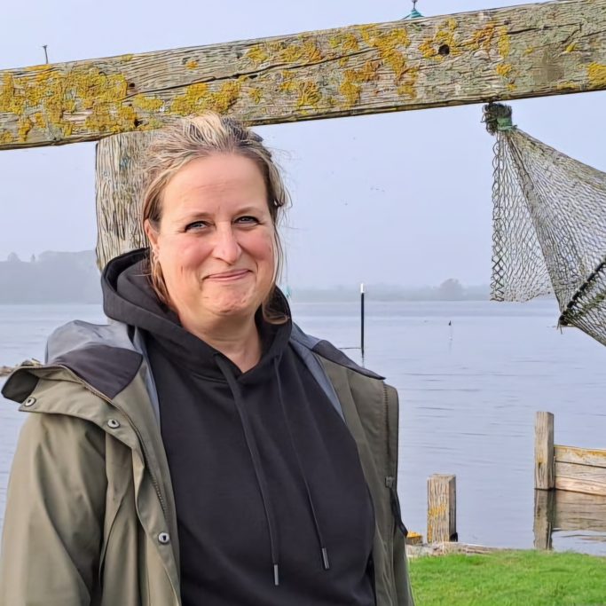 Frau mit Holzsteg im Hintergrund, Wasser und Netzanhänger im Bild.