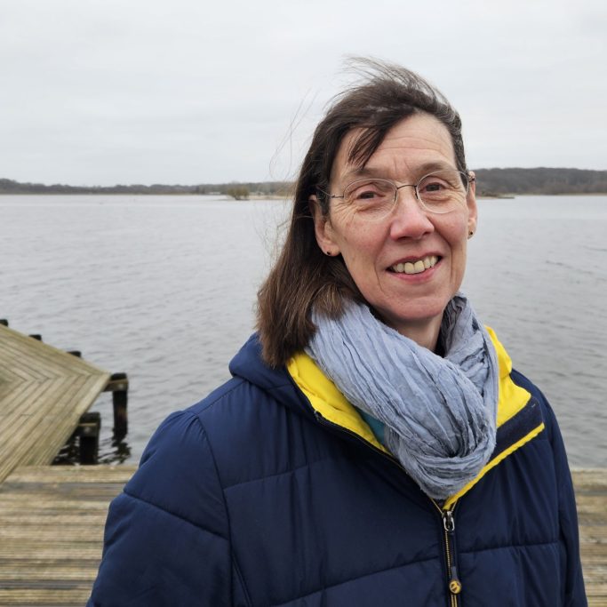 Frau lächelt am Ufer eines ruhigen Sees, umgeben von grauem Himmel.