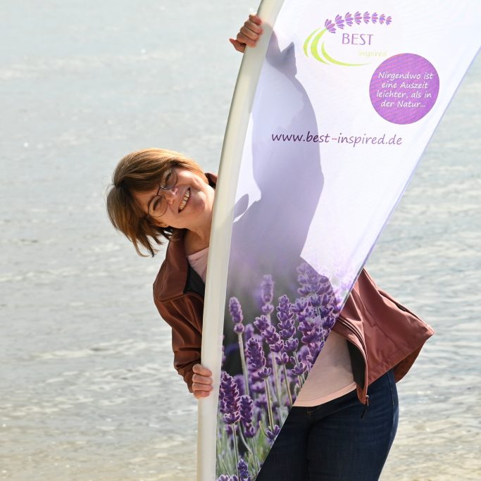 Frau mit einem Banner, umgeben von Lavendel, am Wasser lächelnd.