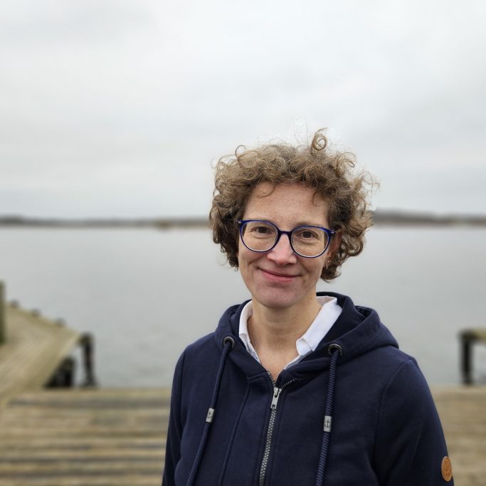 Lächelnde Person mit lockigem Haar und Brille, am Wasser in grauer Umgebung.